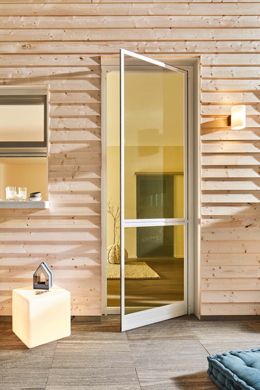 Moderne Sauna aus hellem Holz mit Glastuer und beleuchtetem Wuerfelhocker